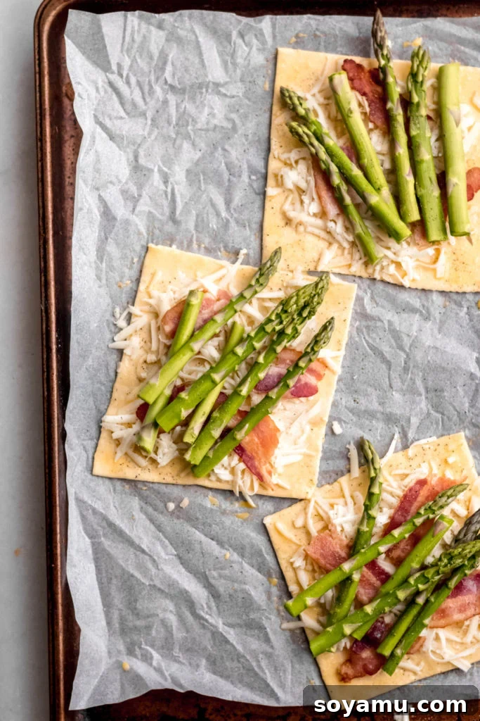 Asparagus Tarts with Bacon and Gruyere - The easiest appetizer or side you'll ever whip up! Made with store-bought puff pastry, fresh asparagus, bacon, and lots of cheese! It's a flavor explosion of sweet, smoky bacon, asparagus, melty cheese, and crisp puff pastry. #asparagus #easterrecipes #asparagus #easyrecipes #easyappetizers #easyeasterrecipes #puffpastry #bacon #gruyere #bluebowlrecipes | bluebowlrecipes.com Asparagus spears arranged on top of puff pastry with bacon and gruyere, ready for baking.