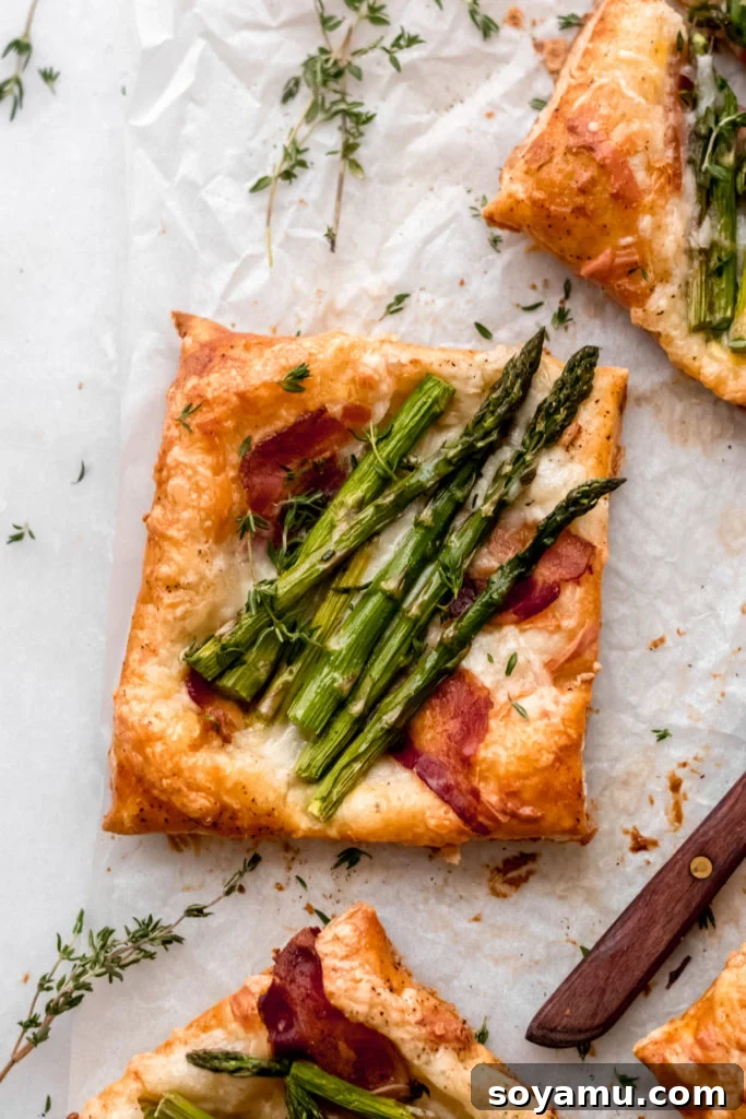 Asparagus Tarts with Bacon and Gruyere - The easiest appetizer or side you'll ever whip up! Made with store-bought puff pastry, fresh asparagus, bacon, and lots of cheese! It's a flavor explosion of sweet, smoky bacon, asparagus, melty cheese, and crisp puff pastry. #asparagus #easterrecipes #asparagus #easyrecipes #easyappetizers #easyeasterrecipes #puffpastry #bacon #gruyere #bluebowlrecipes | bluebowlrecipes.com A close-up view of a single asparagus tart with bacon and gruyere, showing the flaky pastry and vibrant toppings.