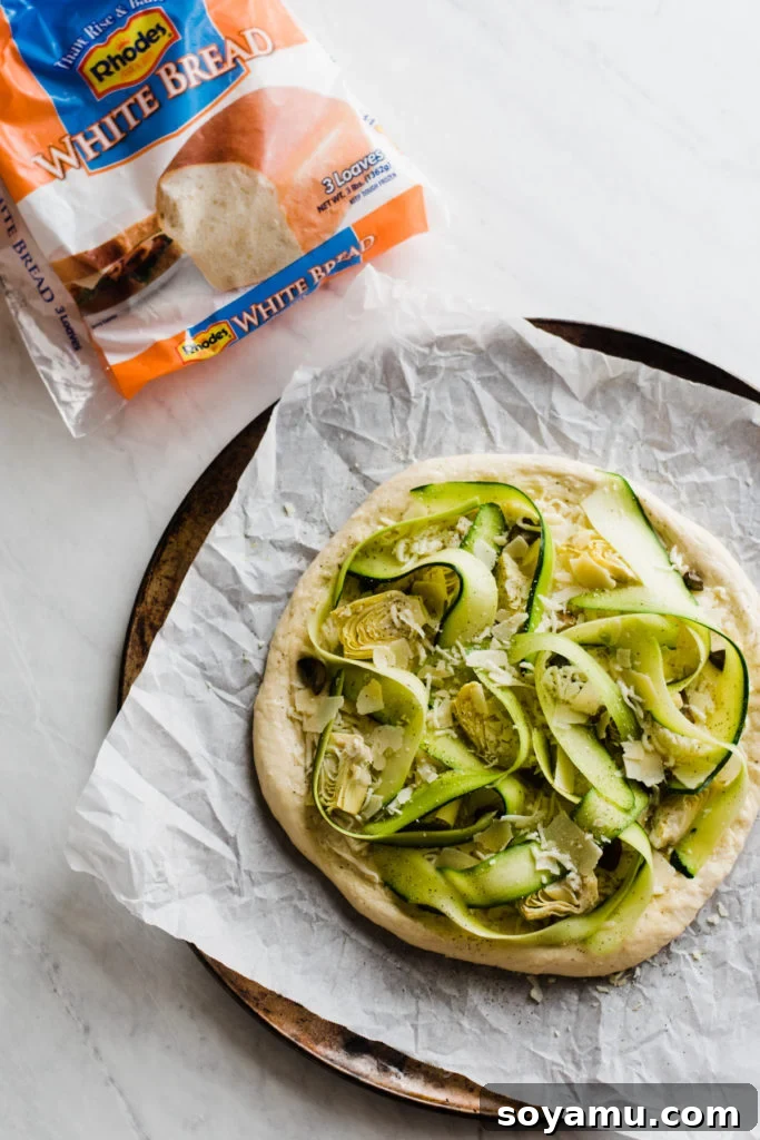 Preparing Green Goddess Pizza with fresh ingredients Raw pizza ingredients layered on top of Rhodes dough, showing artichokes, mushrooms, and zucchini ribbons.