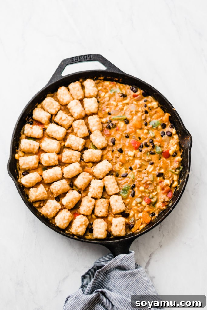 Preparing Tater Tot Hot Dish: Topping with Crispy Tots Half of a cast iron skillet filled with creamy hot dish mixture, with frozen tater tots neatly arranged over the other half, ready for baking.