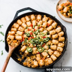 Top down view of tater tot hot dish in a cast iron skillet. There are slices of fresh jalaepenos on top. A wooden spoon is scooping a serving of the casserole.
