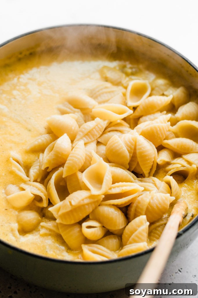 Baked Butternut Squash Mac and Cheese - Incredibly creamy, flavorful, & easy to make! Made with creamy butternut squash sauce, cheddar, parmesan, & smoked gouda cheese, fresh thyme + sage, and a crispy, buttery bread crumb topping. This side will be the star of your Thanksgiving meal! #macandcheese #easyrecipe #vegetarianrecipes #dinnerrecipes #sidedishrecipes #butternutsquashrecipes #thanksgivingrecipes #bluebowlrecipes | bluebowlrecipes.com Close-up of cooked pasta being added to the rich, creamy butternut squash cheese sauce in a Dutch oven.