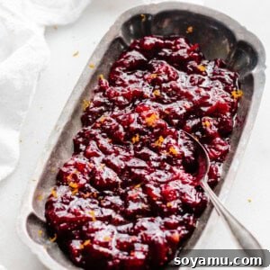 A festive serving dish brimming with homemade cranberry sauce, garnished with vibrant orange zest. A serving spoon gently scoops into the rich, ruby-red sauce.