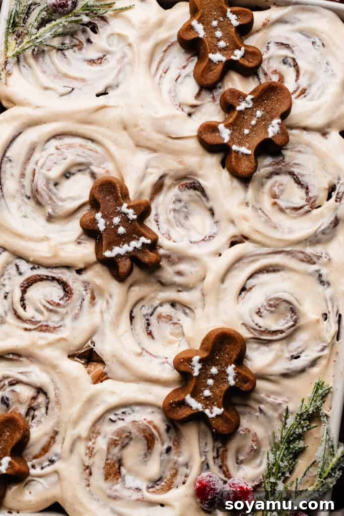 A close-up of frosted gingerbread cinnamon rolls, beautifully decorated with mini gingerbread cookies, sugared rosemary, and cranberries, showcasing festive elegance.