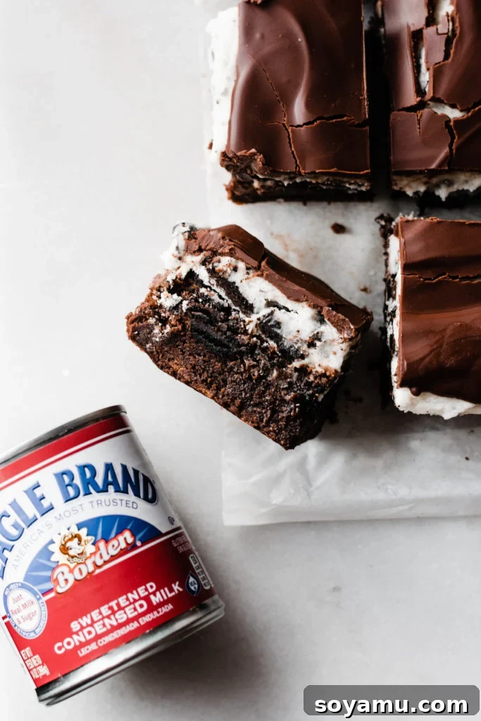 Ultimate Cookies & Cream Bars - The holiday treat to end all holiday treats! Made with a layer of thick, fudgy brownies that have a layer of oreos in the middle, & topped with sweetened condensed milk + crushed oreos. Next up, a layer of oreo frosting and a layer of chocolate to top it all off! This is the stuff of chocolate dreams! #dessertrecipes #christmascookies #brownies #oreo #christmasbaking #christmasrecipes #cookierecipes #frosting #chocolate #bluebowlrecipes | bluebowlrecipes.com A close-up view of the Ultimate Cookies & Cream Bars showcasing their rich, distinct layers: fudgy brownie, whole Oreos, caramelized sweetened condensed milk, crisp crushed Oreos, smooth Oreo frosting, and glossy chocolate ganache. Perfect for festive gatherings and Christmas cookie platters. #dessertrecipes #christmascookies #brownies #oreo #christmasbaking #christmasrecipes #cookierecipes #frosting #chocolate #bluebowlrecipes