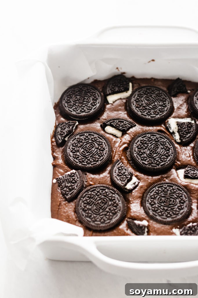 Ultimate Cookies & Cream Bars - The holiday treat to end all holiday treats! Made with a layer of thick, fudgy brownies that have a layer of oreos in the middle, & topped with sweetened condensed milk + crushed oreos. Next up, a layer of oreo frosting and a layer of chocolate to top it all off! This is the stuff of chocolate dreams! #dessertrecipes #christmascookies #brownies #oreo #christmasbaking #christmasrecipes #cookierecipes #frosting #chocolate #bluebowlrecipes | bluebowlrecipes.com Preparing the fudgy brownie base with a layer of whole Oreos for Ultimate Cookies & Cream Bars. This step-by-step image shows the thick brownie batter with a middle layer of chocolate sandwich cookies, essential for a rich homemade brownie dessert. #brownies #oreo #dessertmaking #baking