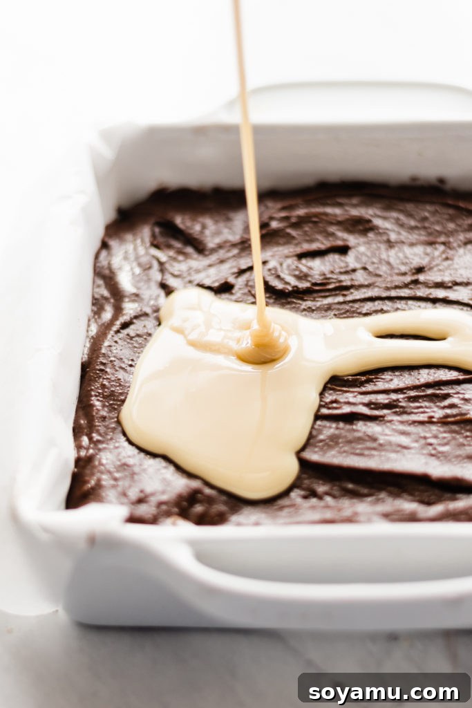 Ultimate Cookies & Cream Bars - The holiday treat to end all holiday treats! Made with a layer of thick, fudgy brownies that have a layer of oreos in the middle, & topped with sweetened condensed milk + crushed oreos. Next up, a layer of oreo frosting and a layer of chocolate to top it all off! This is the stuff of chocolate dreams! #dessertrecipes #christmascookies #brownies #oreo #christmasbaking #christmasrecipes #cookierecipes #frosting #chocolate #bluebowlrecipes | bluebowlrecipes.com Sweetened condensed milk being poured over brownie batter with crushed Oreos for a crunchy topping on Ultimate Cookies & Cream Bars. This image highlights the crucial step of adding a rich, sweet layer before baking, vital for delicious holiday desserts. #sweetenedcondensedmilk #oreocrumble #brownietop