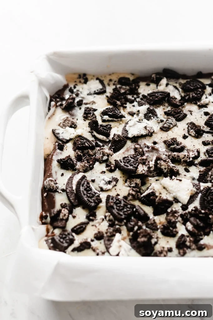 Ultimate Cookies & Cream Bars - The holiday treat to end all holiday treats! Made with a layer of thick, fudgy brownies that have a layer of oreos in the middle, & topped with sweetened condensed milk + crushed oreos. Next up, a layer of oreo frosting and a layer of chocolate to top it all off! This is the stuff of chocolate dreams! #dessertrecipes #christmascookies #brownies #oreo #christmasbaking #christmasrecipes #cookierecipes #frosting #chocolate #bluebowlrecipes | bluebowlrecipes.com Crushed Oreos generously sprinkled over sweetened condensed milk on brownie batter, showing the bars ready for the oven. This visual emphasizes the final preparation step before baking the Ultimate Cookies & Cream Bars, ensuring a delightful texture in every bite. #oreotopping #browniesreadyforoven