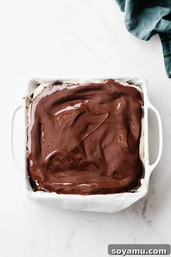 Ultimate Cookies & Cream Bars - The holiday treat to end all holiday treats! Made with a layer of thick, fudgy brownies that have a layer of oreos in the middle, & topped with sweetened condensed milk + crushed oreos. Next up, a layer of oreo frosting and a layer of chocolate to top it all off! This is the stuff of chocolate dreams! #dessertrecipes #christmascookies #brownies #oreo #christmasbaking #christmasrecipes #cookierecipes #frosting #chocolate #bluebowlrecipes | bluebowlrecipes.com Smooth chocolate ganache being poured over Oreo frosting, preparing Ultimate Cookies & Cream Bars for chilling. This final, decadent chocolate layer creates a glossy finish for festive desserts and enhances the overall rich flavor profile. #chocolategnache #dessertbars #holidaydesserts