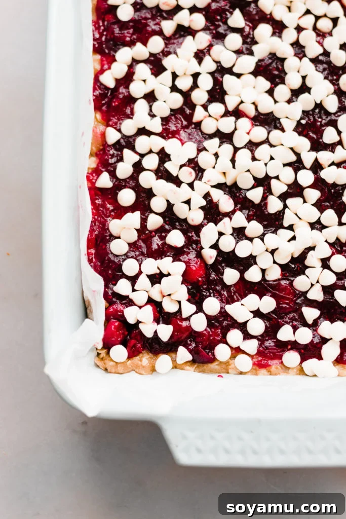 The process of layering Cranberry White Chocolate Crumble Bars, showing the cranberry sauce spread over the base, topped with an abundance of white chocolate chips.
