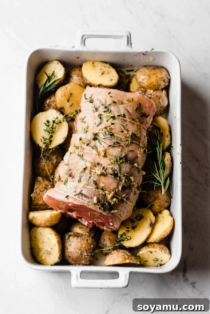 The lamb roast and potatoes roasting together in the oven. Lamb Roast with Herbed Garlic Butter Potatoes roasting in oven - A whole lamb leg placed fat-side up over a bed of herbed garlic butter potatoes, with fresh rosemary and thyme sprigs, all roasting in a hot oven. Captures the one-pan cooking method for this delicious holiday dinner. #RoastingLamb #OvenBaked #ComfortFood #DinnerIdeas #FoodPhotography