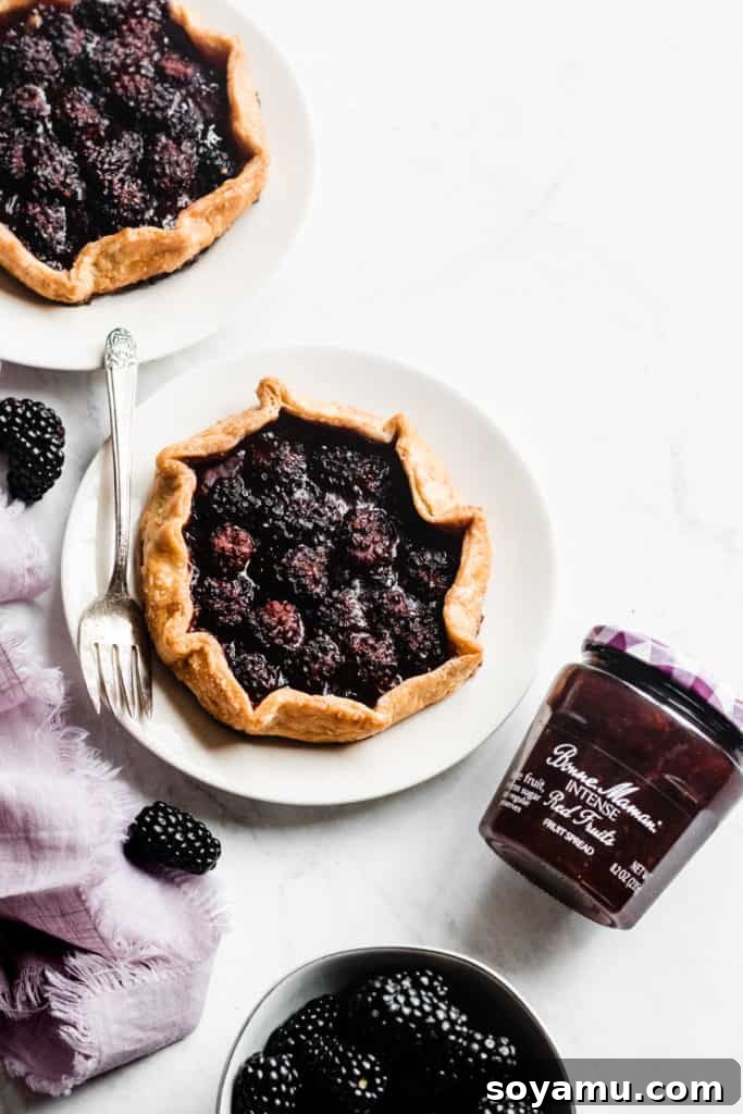 A close-up shot of a single baked mini galette, highlighting its golden, rustic whole wheat crust and the rich, dark red fruit filling peeking through. Glistening fresh blackberries are artfully placed on top, adding to its appeal.