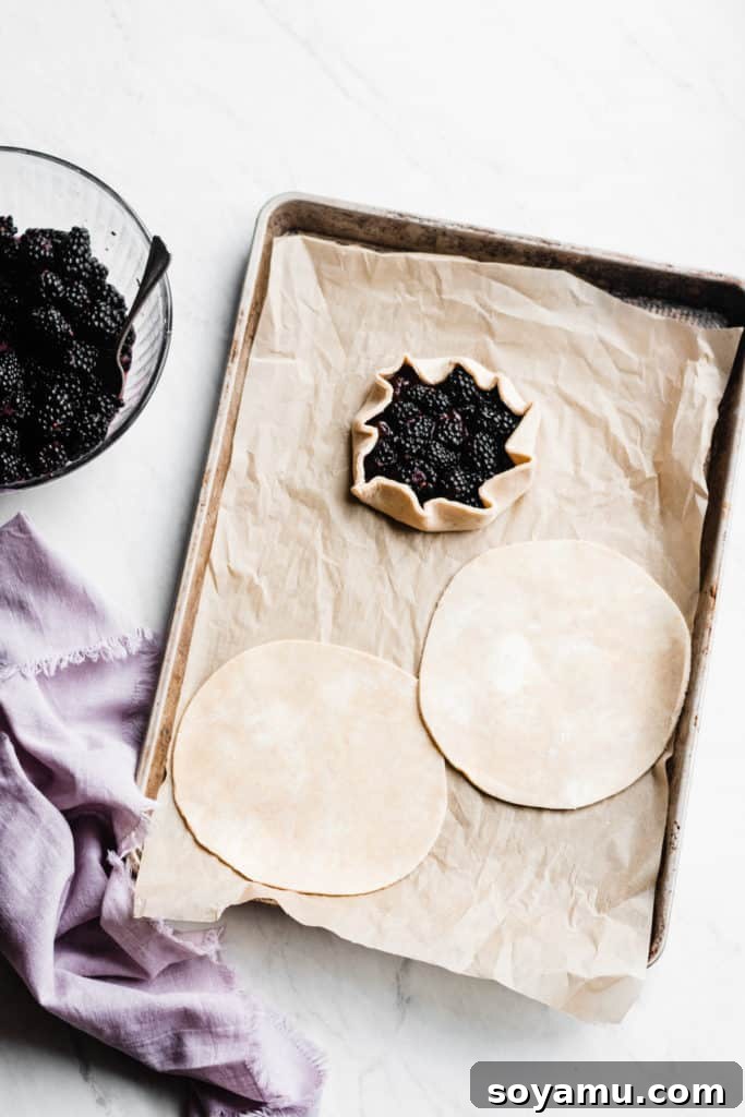 A pair of hands gently spreading a rich, dark red Bonne Maman INTENSE Red Fruits Fruit Spread onto a circular piece of raw whole wheat galette dough on parchment paper. The rustic edges of the dough are visible.