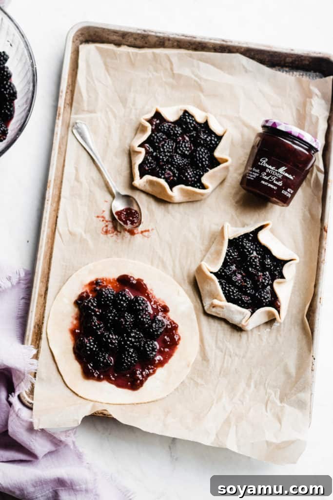 A close-up of an uncooked mini galette on parchment paper, generously filled with a thick layer of red fruit spread and whole, plump fresh blackberries in the center. The dough edges are still unfolded, ready for the next step.