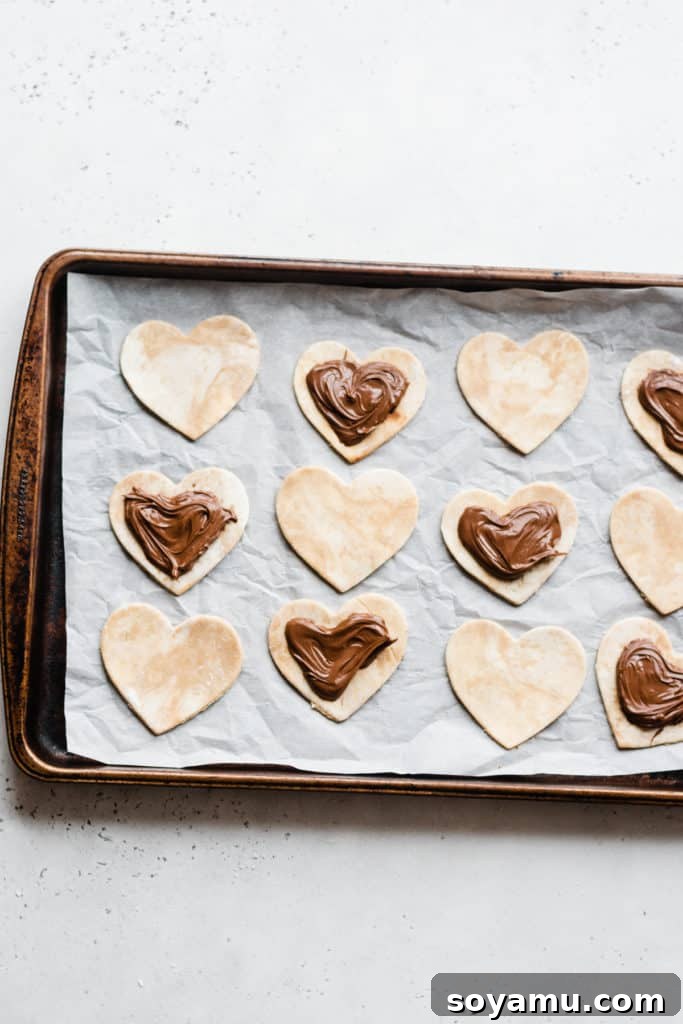 Nutella filling on dough hearts Heart-shaped pie dough bases, each topped with a generous dollop of Nutella, ready for the top layer and sealing.