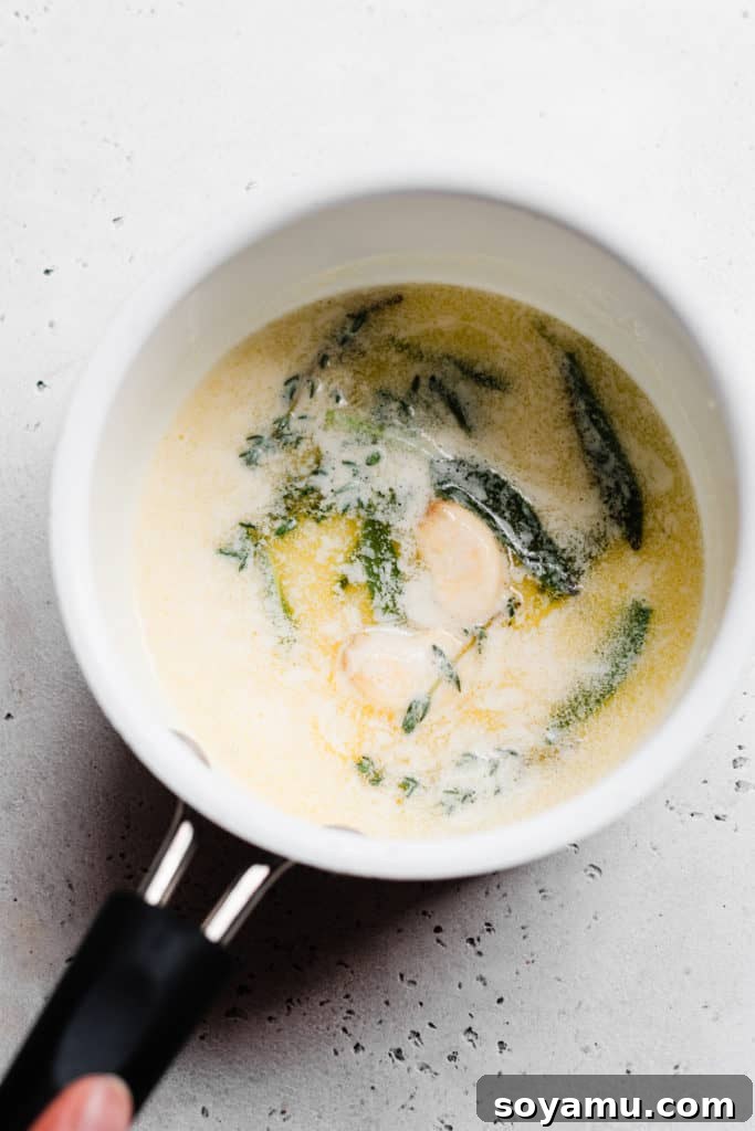 Infusing Butter with Garlic and Herbs for Mashed Potatoes. A small saucepan containing melted butter, a smashed garlic clove, and fresh herbs (sage and thyme), infusing on low heat.