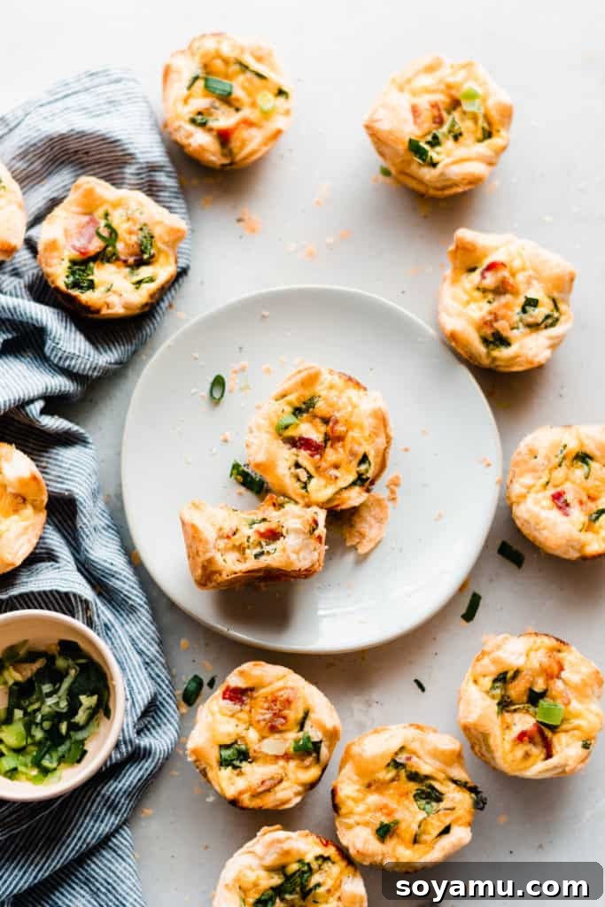 Mini Quiche Cups with Spinach, Bacon, and Cheddar - Made with store-bought puff pastry, filled with a delicious smoky, flavorful mixture of gruyere and cheddar cheese, spinach, bacon, eggs, and milk. Perfect for Easter brunch. #easterrecipes #brunchrecipes #quiche #breakfastrecipes #brunch #easterbrunch #spinachquiche #bacon #miniquiche A half-eaten mini quiche cup on a plate, surrounded by more perfect mini quiches, ready for serving.