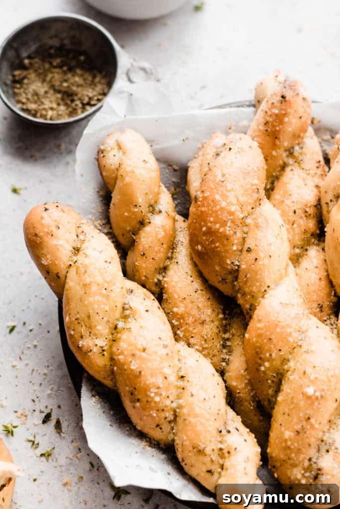 Close up of a plate full of buttery twisted breadsticks sprinkled with parmesan and herbs, showcasing their texture and golden finish.