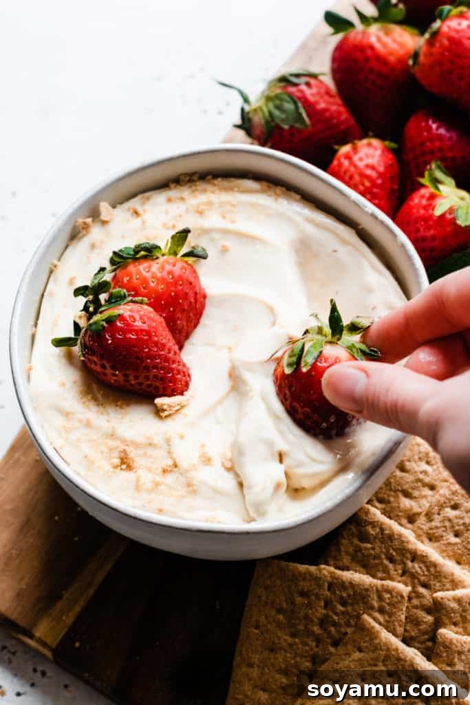 Creamy 4-Ingredient Cheesecake Dip - The perfect no-bake dessert for any occasion, easy enough for kids to help! Serve with fresh fruit and graham crackers for a stunning dessert board. A hand dipping a vibrant red strawberry into a smooth, creamy bowl of cheesecake dip, showcasing the perfect texture and inviting presentation.