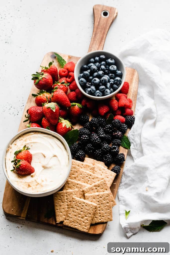 Versatile Cheesecake Dip Dessert Board - Perfect for parties, holidays, or any time you need a quick, delicious no-bake treat that caters to all tastes. A beautifully arranged wooden board with a central bowl of creamy cheesecake dip, surrounded by an inviting selection of golden graham crackers, fresh red strawberries, and plump blueberries, all ready for dipping.