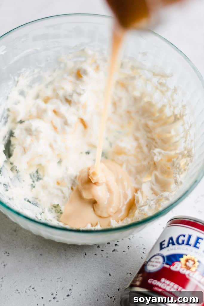 Cheesecake Dip Ingredient Prep - Combining softened cream cheese with sweetened condensed milk for a smooth and creamy base. Sweetened condensed milk being poured gracefully into a bowl of perfectly softened cream cheese, signifying the beginning of the mixing process for the dip.
