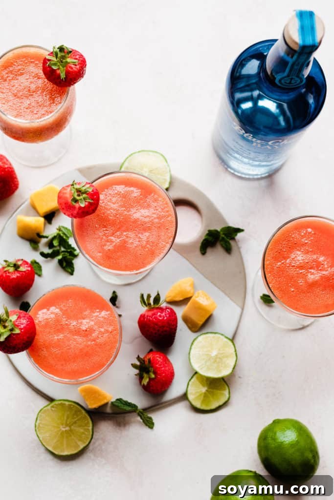 Three cocktail glasses filled with a bright pinky orange rum punch, with mangos, strawberries, and lime slices scattered around, and a blue rum bottle