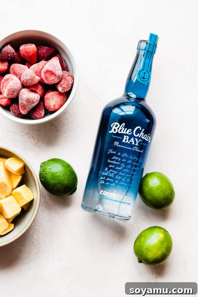 A bowl of frozen strawberries, frozen mango, and a few limes scattered around a blue Blue Chair Bay Rum Coconut Rum bottle
