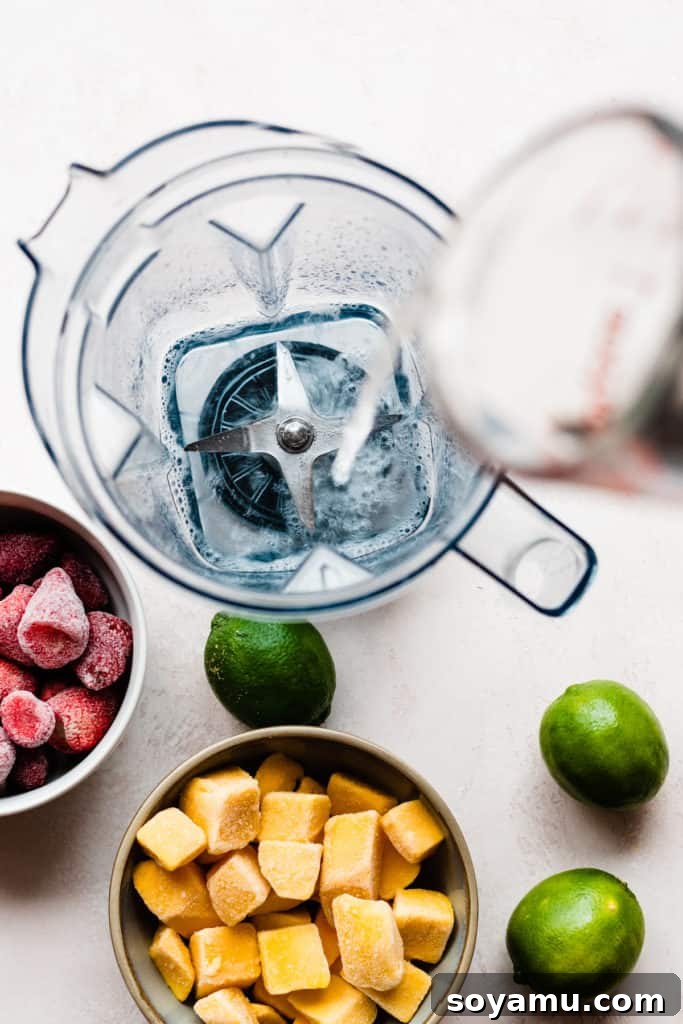 Coconut rum being poured into a blender, with strawberries, mango, and limes scattered around