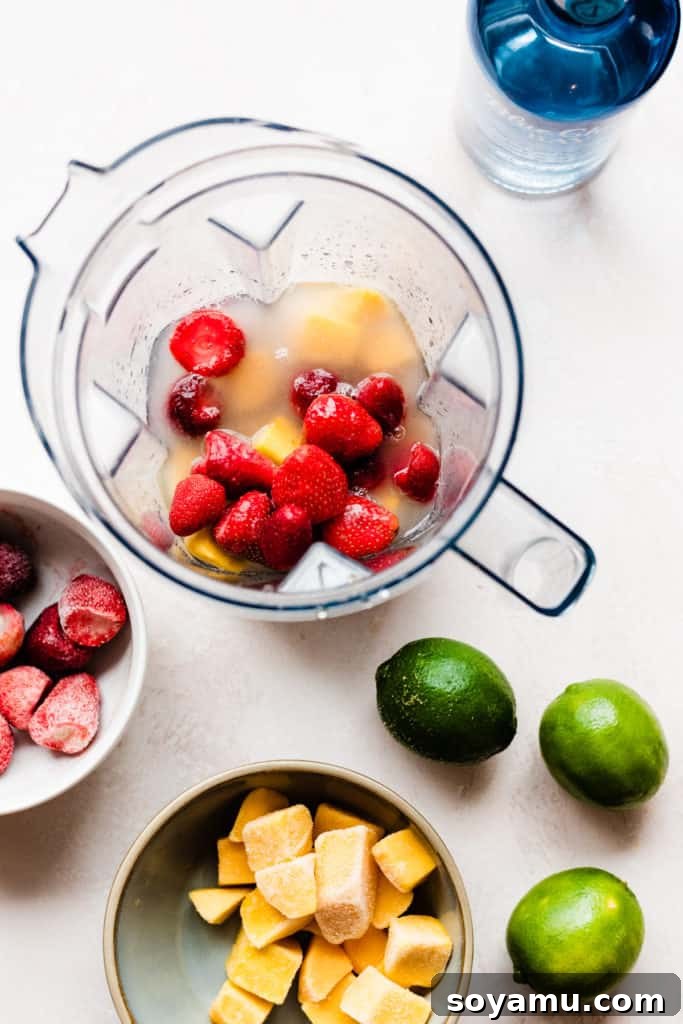 A blender filled with frozen strawberries and mango pieces, and coconut rum, surrounded by pieces of the fruit