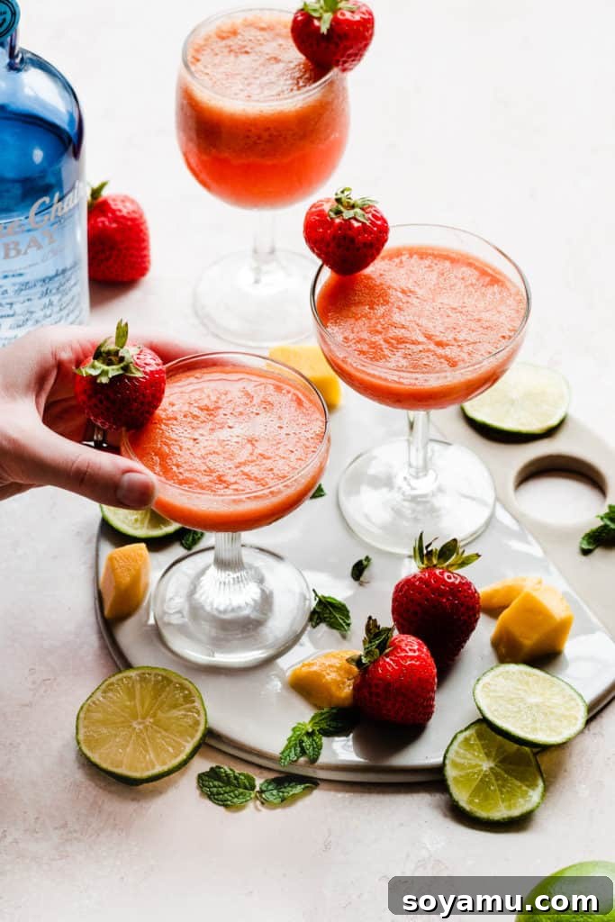Three cocktail glasses filled with a bright pinky orange frozen punch, and garnished with fresh strawberries, with fruit scattered around, and a hand picking up one of the glasses