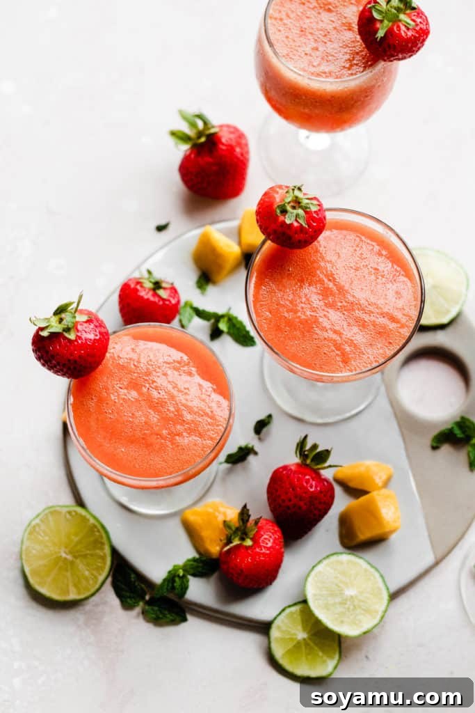 Three cocktail glasses from above, filled with a bright pinky orange punch and topped with fresh strawberries, with lime slices, mango chunks, and strawberries scattered around