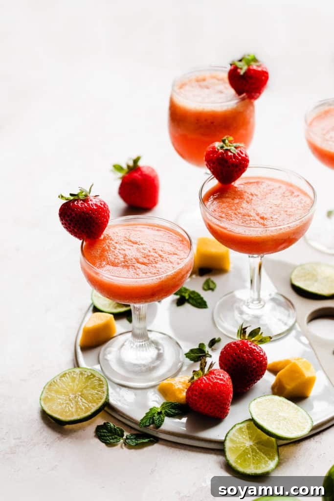 Four cocktail glasses filled with a bright pinky orange rum punch, with mangos, strawberries, and lime slices scattered around
