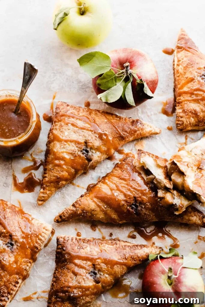 Flaky Caramel Apple Pockets 2 Puff pastry apple turnovers drizzled with salted caramel sauce on a brown surface with whole apples sitting around.