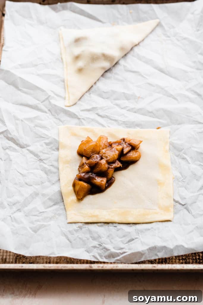 Flaky Caramel Apple Pockets 6 A square of puff pastry, with egg wash brushed over the edges, and apple filling on one half. A folded apple turnover is in the background on the same baking sheet