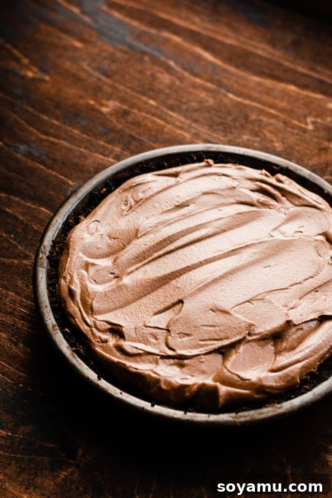 A chocolate cookie crust filled with creamy chocolate mousse, ready for the next layer, sits in a metal pie plate on a wooden tabletop.