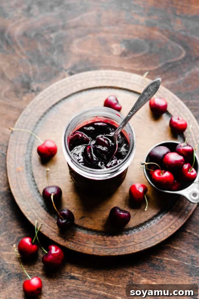 A glass jar filled with luscious, homemade boozy cherry sauce, surrounded by a scattering of fresh, ripe cherries on a wooden table, hinting at its deliciousness.