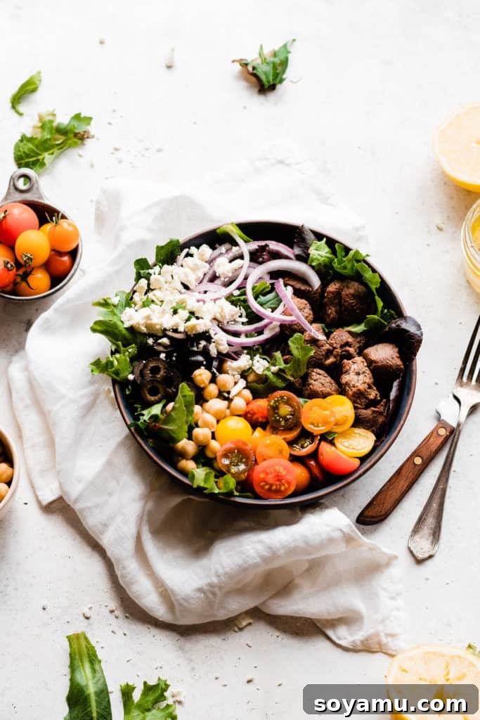 Zesty Lemon Grilled Lamb Power Bowls 2 A vibrant bowl filled with lettuce, crumbled feta, thinly sliced red onion, black olives, halved cherry tomatoes, and perfectly grilled lamb meat, all resting on a smooth surface with a white linen and fresh lemon slices scattered around