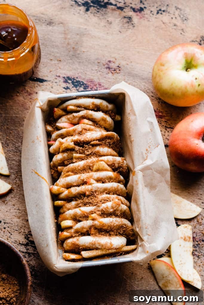 Gooey Caramel Apple Monkey Bread 4 A loaf pan meticulously lined with parchment paper, showcasing layers of dough, vibrant apple slices, rich caramel sauce, and a generous sprinkle of brown sugar and cinnamon, all prepped for baking.
