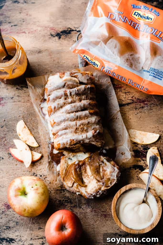 Gooey Caramel Apple Monkey Bread 6 The fully glazed Caramel Apple Pull Apart Bread sits on a wooden surface, surrounded by fresh apples, a jar of salted caramel, and the signature orange bag of Rhodes dinner rolls, highlighting the convenience of the pre-made dough.