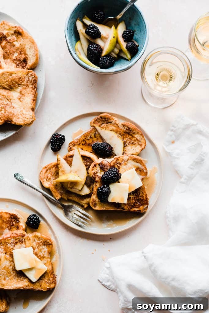 Indulgent Vanilla Rum French Toast with Blackberries and Pears 2 Two plates of golden french toast topped with butter, maple syrup, and blackberries and pear slices, on a light pink surface with two glasses of apple juice and a white linen