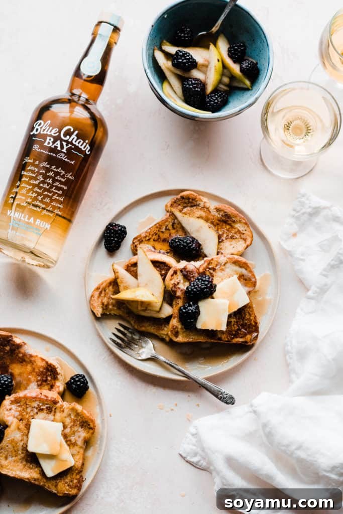 Indulgent Vanilla Rum French Toast with Blackberries and Pears 6 Two plates of golden french toast topped with butter, maple syrup, and blackberries and pear slices, on a light pink surface with two glasses of apple juice and a white linen. A bottle of Blue Chair Bay Vanilla Rum is laying next to the plates