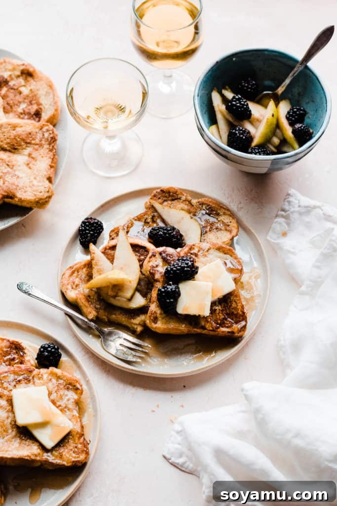 Indulgent Vanilla Rum French Toast with Blackberries and Pears 7 Two plates of golden french toast topped with butter, maple syrup, and blackberries and pear slices, on a light pink surface with two glasses of apple juice and a white linen. A bite has been taken from the french toast