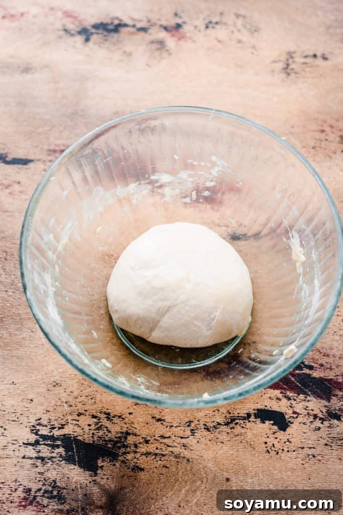 Decadent Deep Dish Pear and Cinnamon Crumble Pizza 3 A glass bowl with unrisen pizza dough on a wooden surface, showing the initial stage of dough preparation