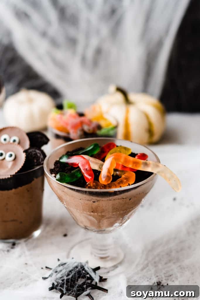 A dirt cup topped with oreo dirt and gummy worms, with spider webs and pumpkins in the background