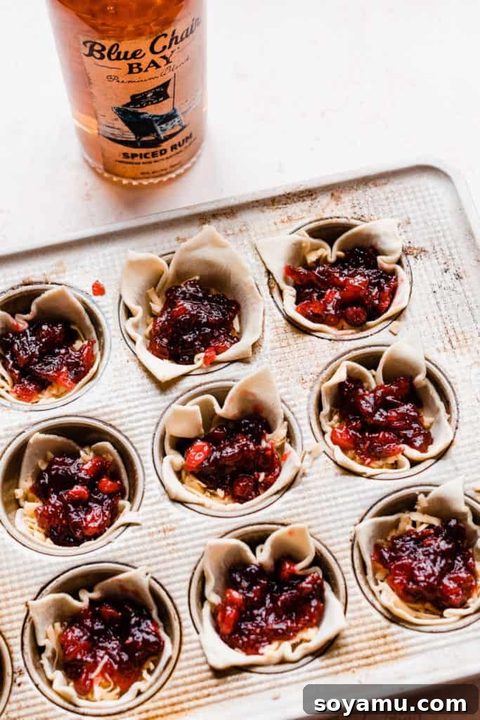 Spiced Rum Cranberry and Gouda Puff Pastry Bites 6 A muffin tin filled with unbaked puff pastry bites, each containing gouda cheese and cranberry sauce, with a bottle of spiced rum blurred in the background, hinting at the key ingredient.