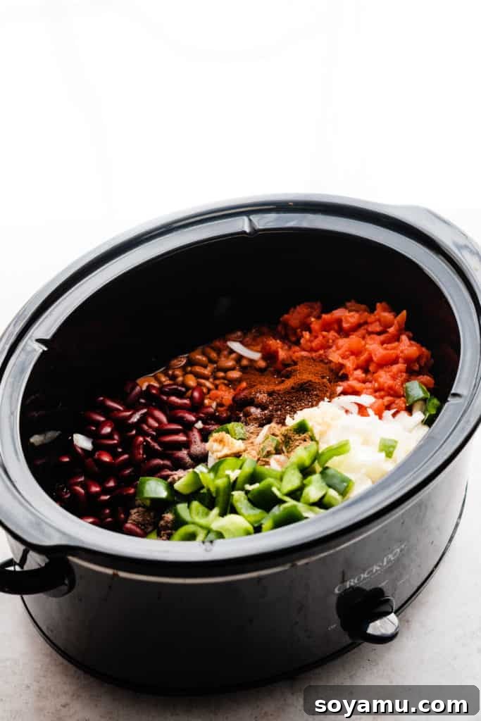 Effortless Slow Cooker Chili Perfection 4 A slow cooker pot brimming with all the raw ingredients for chili: ground beef, diced onions and bell peppers, canned tomatoes, and beans, before being stirred or cooked.