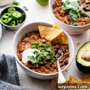 Two white bowls of slow cooker chili with meat and beans. The bowls are topped with jalapeno slices, fresh cilantro, and a scoop of sour cream. Bowl closest to camera has a tortilla chip and spoon in the bowl.