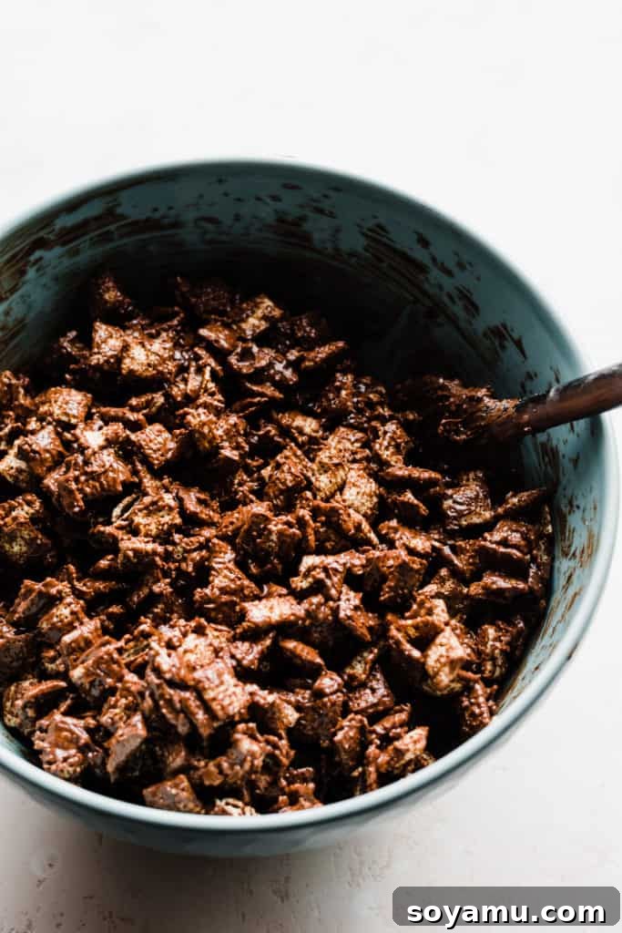 A large mixing bowl filled with crispy Chex cereal, perfectly coated in a rich, glossy chocolate and peanut butter mixture. The cereal glistens, indicating it's thoroughly enrobed and ready for the crucial next step of being dusted with powdered sugar.
