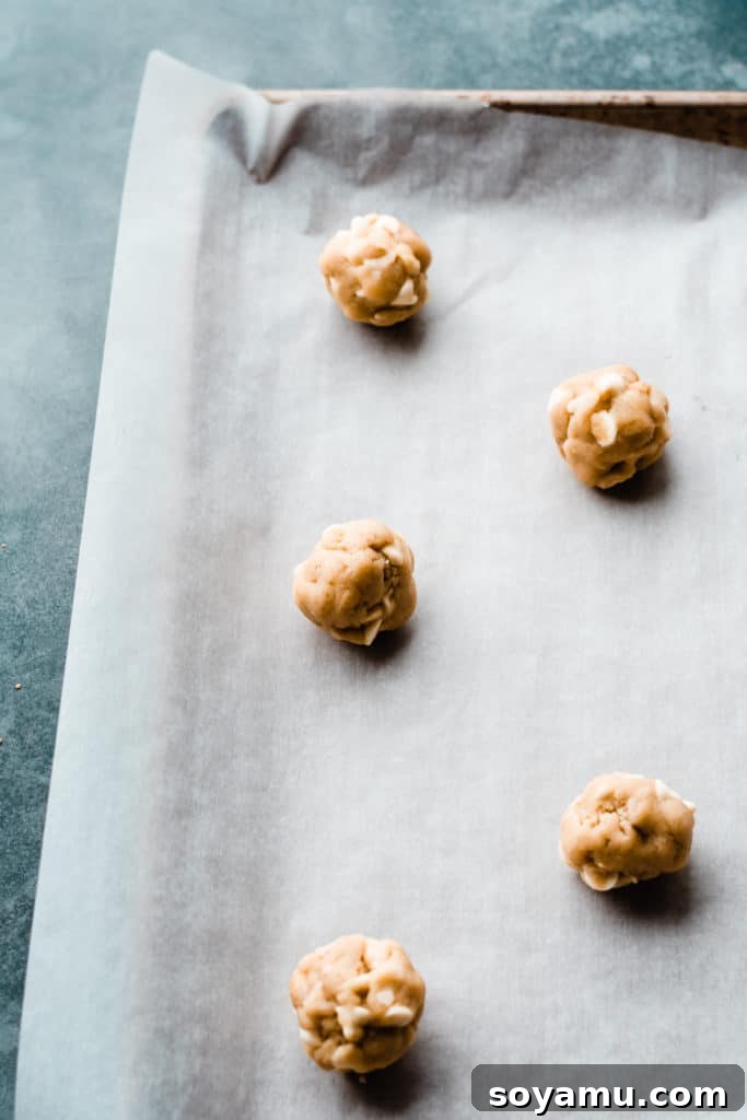 Portioned cookie dough balls, generously studded with white chocolate chips and macadamia nuts, neatly arranged on a parchment-lined baking sheet, awaiting their turn in the oven.