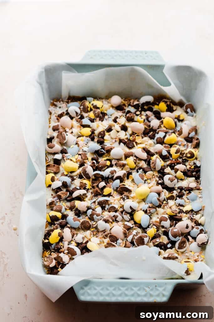 A pan of meticulously assembled Seven Layer Bars, ready for baking, topped with an array of chocolate chips and festive Easter candies.
